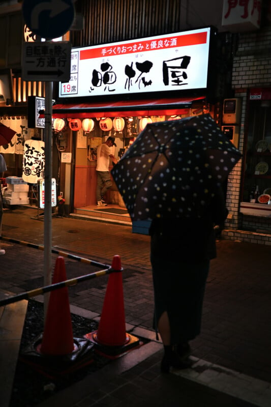 A person with a polka dot umbrella walks towards a brightly lit Japanese restaurant entrance at night. Red lanterns hang above the entrance, and an illuminated sign features Japanese text. Wet pavement and traffic cones are visible in the foreground.