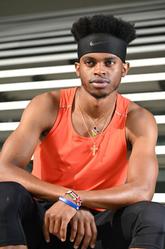 A person wearing an orange tank top, black headband, and several necklaces and bracelets, sits on steps. They have short hair and are looking at the camera with a neutral expression.