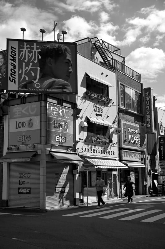 A black and white photo of a city street corner featuring a multi-story building with various signs. Two pedestrians cross the street in front of a "Bakery & Burger" shop. A large billboard with a person's face is visible on the building.