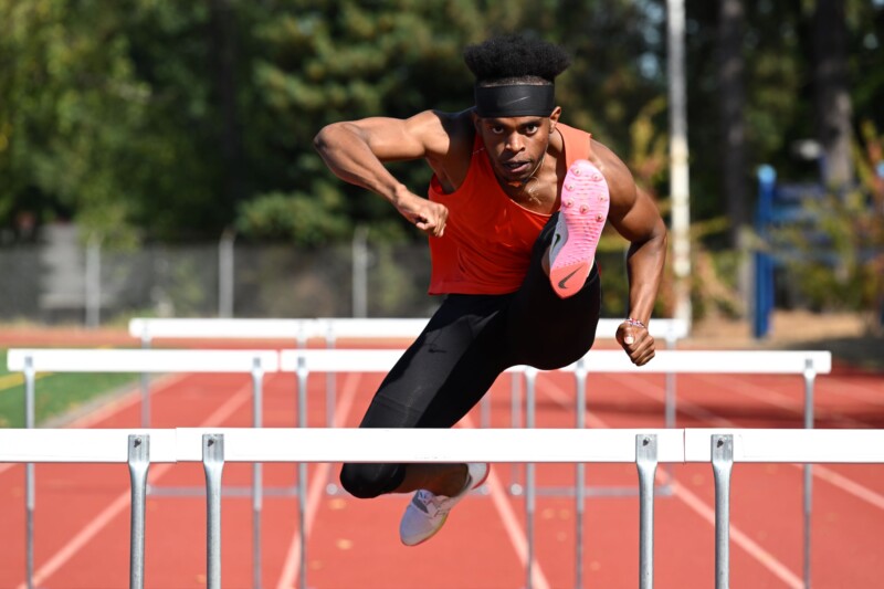 A runner in a red tank top and black leggings is mid-air, clearing a hurdle on an outdoor track. His expression is focused, and he wears a headband. The background shows trees and a sunny day.