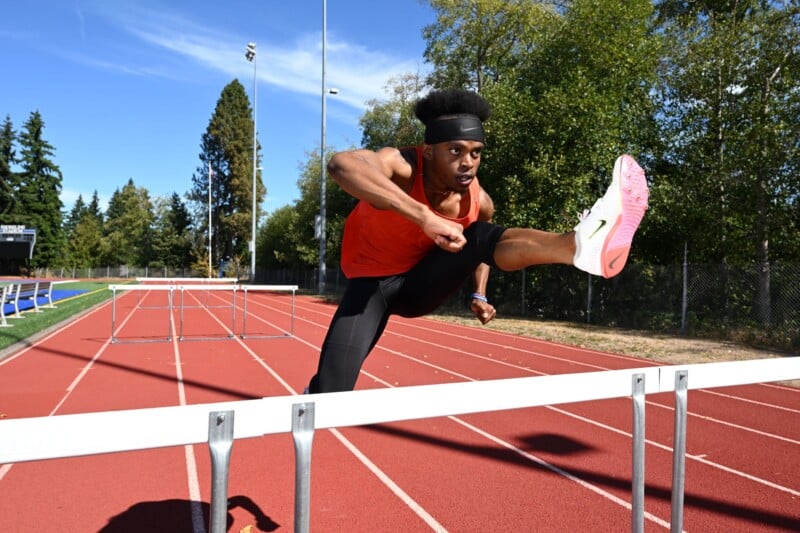 An athlete in an orange tank top and black leggings is mid-jump over a hurdle on a red track. Trees and a clear blue sky are in the background.