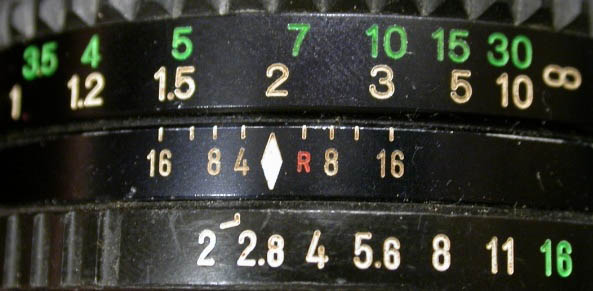 Close-up of a camera lens with focus and aperture markings. The upper ring shows distance scale numbers in green and white. Below, the aperture ring displays f-stop numbers in white and red, ranging from 2.8 to 16.