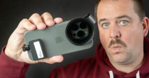 A person with a mustache holds a smartphone with a large lens attachment in front of their face. They are wearing a maroon hoodie and making a surprised expression against a plain dark background.