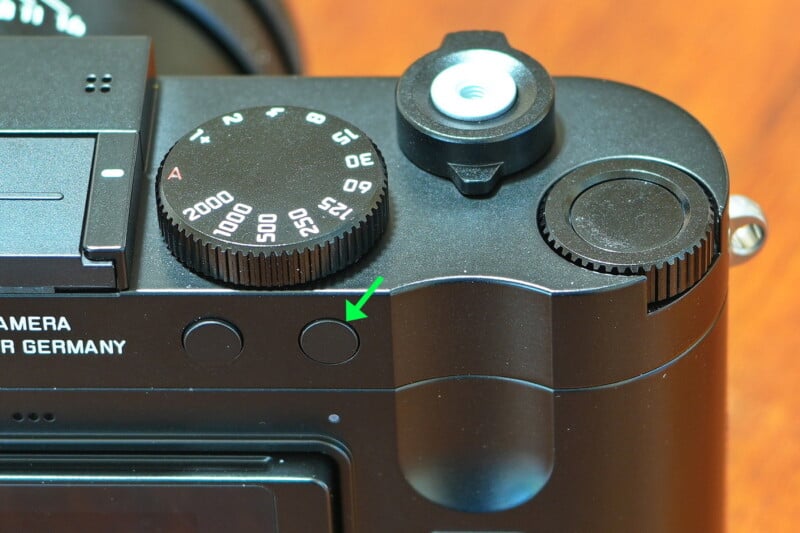 Close-up of a black camera with a focus on its settings dial, featuring shutter speeds from 60 to 2000. A green arrow points to a button next to the dial. The camera is labeled "MADE IN GERMANY.