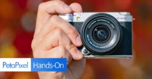 A person holding a Fujifilm X-M5 camera with an aspherical lens against a blurred background of red and orange hues. The image includes the PetaPixel logo and the text "Hands-On" at the bottom left.