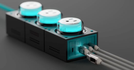 A sleek, modern black power strip with three turquoise-lit outlets. Multiple gray braided cables are plugged into the side, and the strip has a futuristic design with ventilation holes. The background is a dark gray surface.