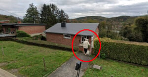 A person circled in red is walking towards the back door of a small brick house surrounded by hedges. The scene is set on an overcast day with trees and a hilly landscape in the background.