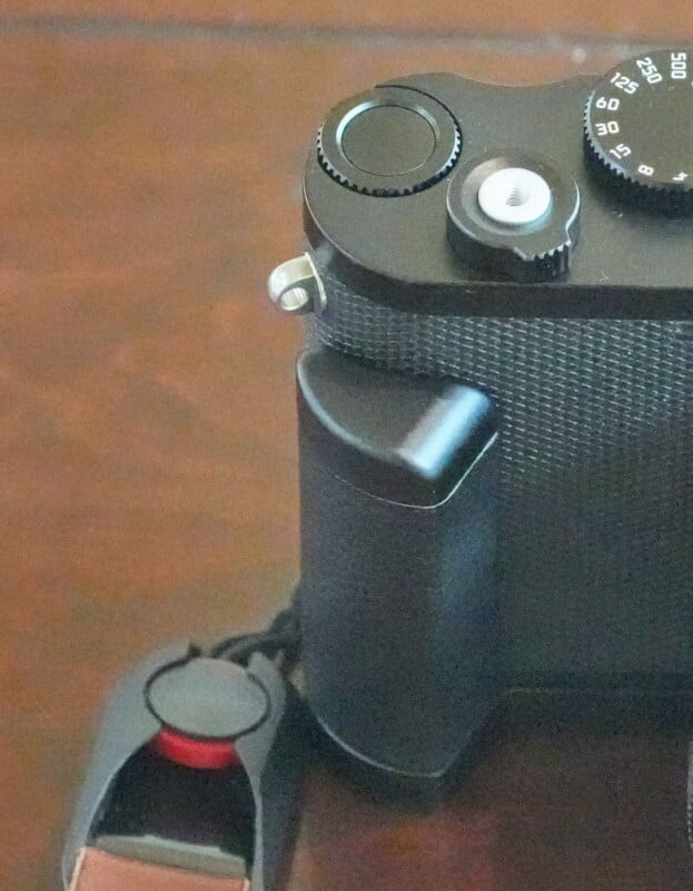 Close-up of a black camera on a wooden surface, showing the top dials and buttons along with the grip handle. A camera strap with a red button detail is visible on the bottom left.