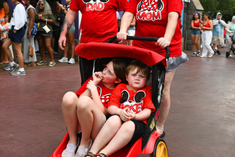 A group of people wearing matching red shirts with mouse ears and family titles. Two children are seated in a red and black stroller. Crowds are visible in the background as they walk along a pathway.