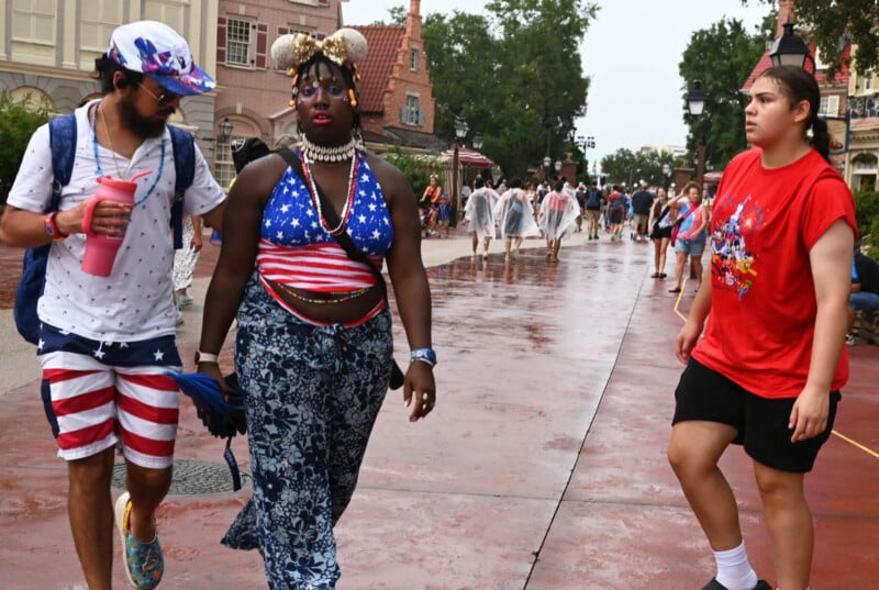 Three people walk on a wet street, surrounded by others in rain ponchos. The person in the middle wears a star-spangled outfit, while the others are in casual clothing. Buildings with varied architecture line the background.