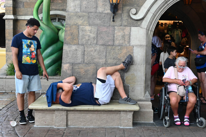 A young man in a Mickey Mouse shirt stands next to an adult resting on a bench. A woman in a wheelchair holds a water bottle nearby. People are exiting a building with an entrance resembling a castle wall.