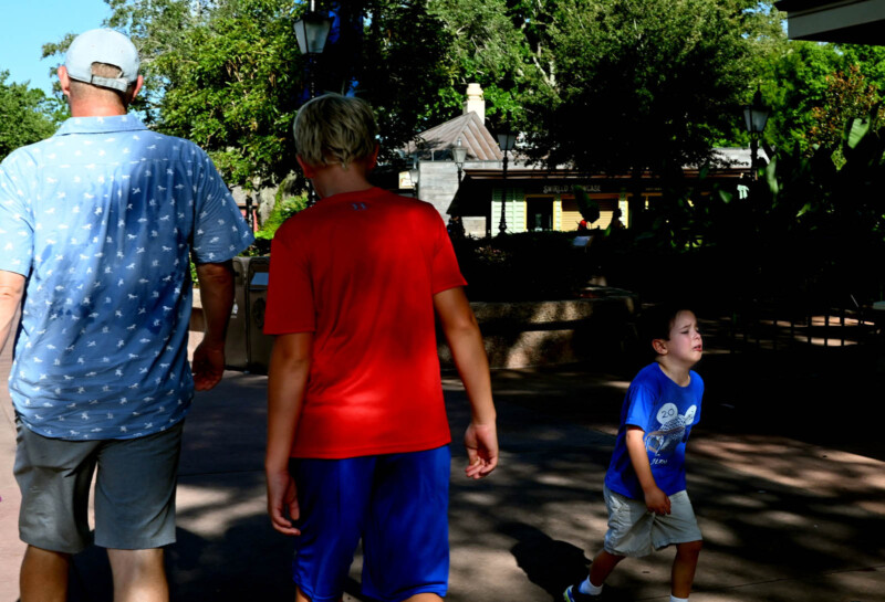 Three people walking along a pathway in a sunny outdoor setting. An adult in a blue shirt and cap is accompanied by two children, one in a red shirt and shorts, the other in a blue shirt and shorts. Trees and a building are visible in the background.