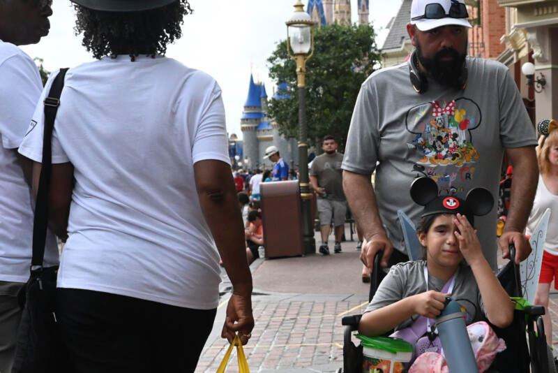 A young girl wearing Mickey Mouse ears sits in a wheelchair pushed by a man in a Disney-themed shirt. They are in an amusement park, surrounded by people and near a castle structure.