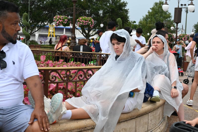 People wearing rain ponchos sit on a stone ledge at a theme park. A man in a white shirt sits nearby, while others are seen walking in the background. The area is decorated with flowers and greenery.