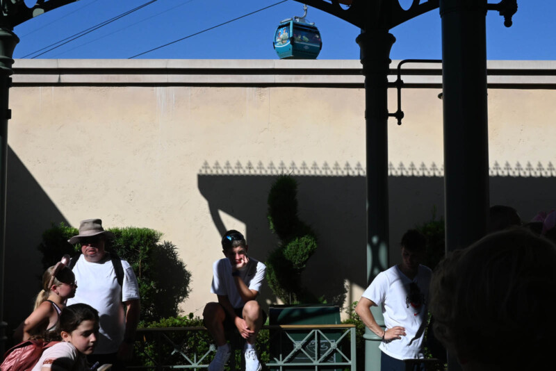 A group of people stands under a shaded area, with two men leaning on a railing. In the background, a gondola car is suspended on a cable against a blue sky. Shadows and silhouettes are cast by the structure overhead.