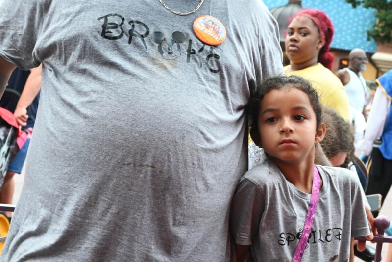 A man wearing a "Broke" shirt with a Disney-themed button stands next to a young girl in a "Spoiled" shirt. They are surrounded by people, including a woman with red hair. The girl looks slightly away, leaning against the man.