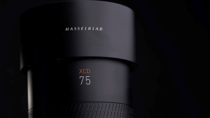 Close-up of a black Hasselblad camera lens with the brand name engraved on the upper cylinder. The text "XCD 75" is printed in orange on the lower part of the lens, set against a dark background.