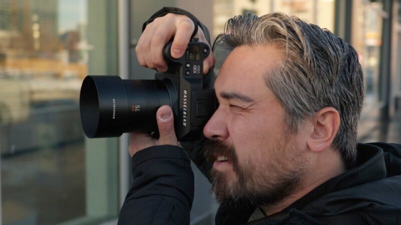 A man with gray hair and a beard is holding a camera up to his eye, taking a photo. He is standing outdoors in front of a building, wearing a dark jacket, with a focused expression.