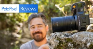 A camera with a large lens is perched on a rock outdoors. In the background, a person with a beard and mustache is slightly out of focus, smiling and looking at the camera. The text "PetaPixel Reviews" is displayed in the upper left corner.