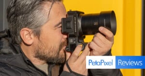 A person with gray hair is using a Hasselblad camera. They are wearing a black jacket and focusing intently, standing next to a yellow wall. The image has a "PetaPixel Reviews" overlay at the bottom right.