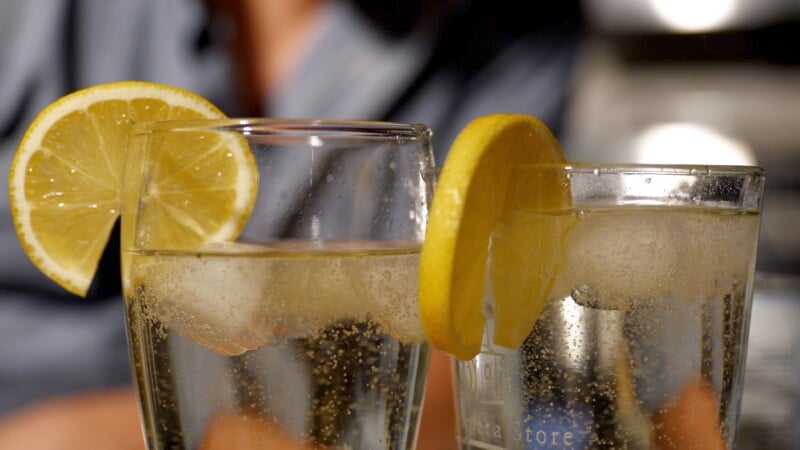 Two glasses of sparkling water with ice cubes, each garnished with a lemon slice, on a blurred background. Sunlight highlights the bubbles and lemons, creating a refreshing and summery atmosphere.