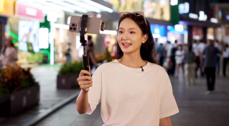 A person in a white t-shirt is smiling while holding a smartphone on a stabilizer, recording or streaming in a vibrant, bustling street at night, illuminated by bright lights and colorful storefronts.