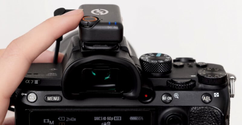 A person's hand adjusts a wireless device attached to the top of a digital camera, focusing on the camera's viewfinder and controls. The device has a small button and indicator lights.