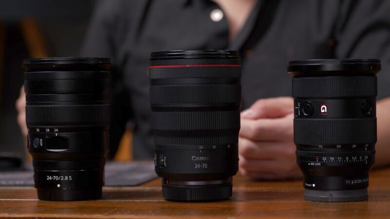 Three camera lenses are placed on a wooden surface. From left to right, they are labeled: 24-70 2.8 S, Canon 24-70, and 24-70 GM. A person in a dark shirt is visible in the background, out of focus.