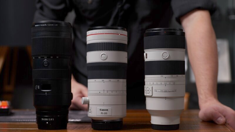 Three camera lenses, including Canon models, stand on a wooden table. A person in a dark shirt is visible in the background, leaning forward with arms on the table.