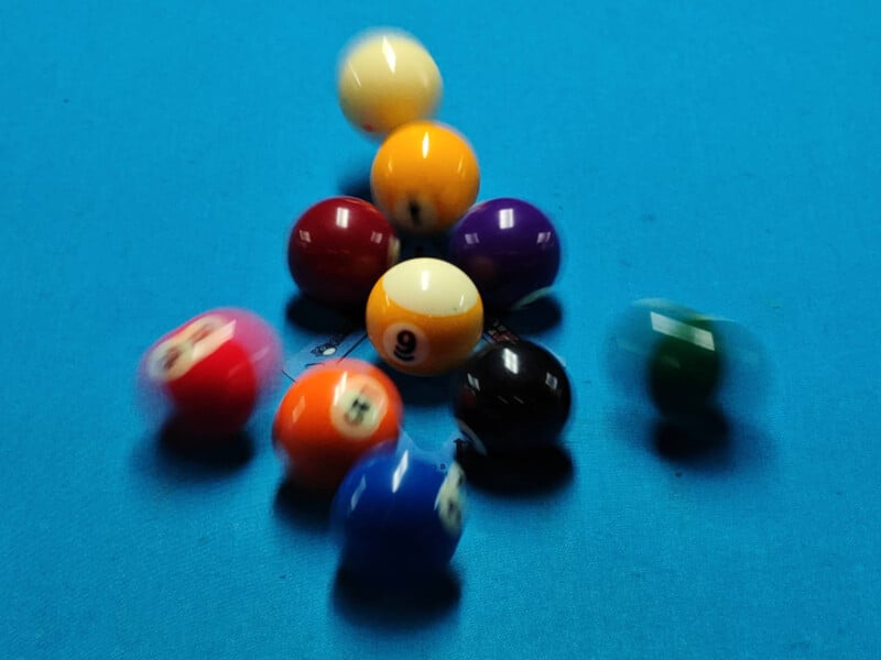 Billiard balls in motion on a blue pool table, captured mid-break. The balls are various colors, creating a dynamic and blurred effect as they scatter across the table.