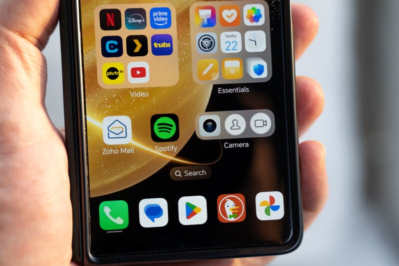 Close-up of a smartphone screen held in hand, displaying various app icons like Spotify, Camera, and a search bar at the bottom. The background features a golden, swirling design.