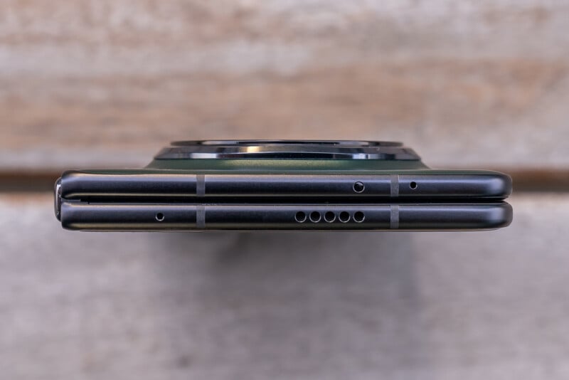 Close-up of two stacked smartphones viewed from the top edge. Both devices are black with visible side buttons and port openings. The background is a blurred wooden surface.