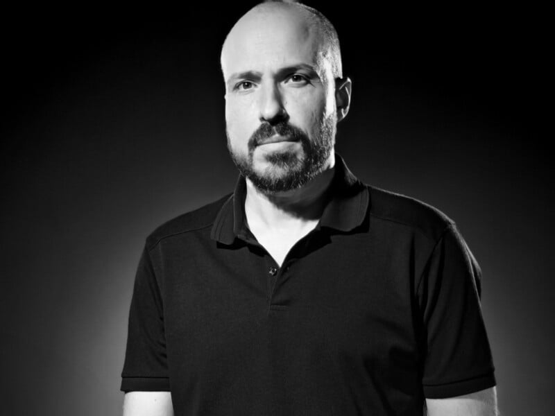 A black and white portrait of a man with a shaved head and beard, wearing a dark polo shirt. He has a serious expression and is set against a dark background, with light highlighting one side.