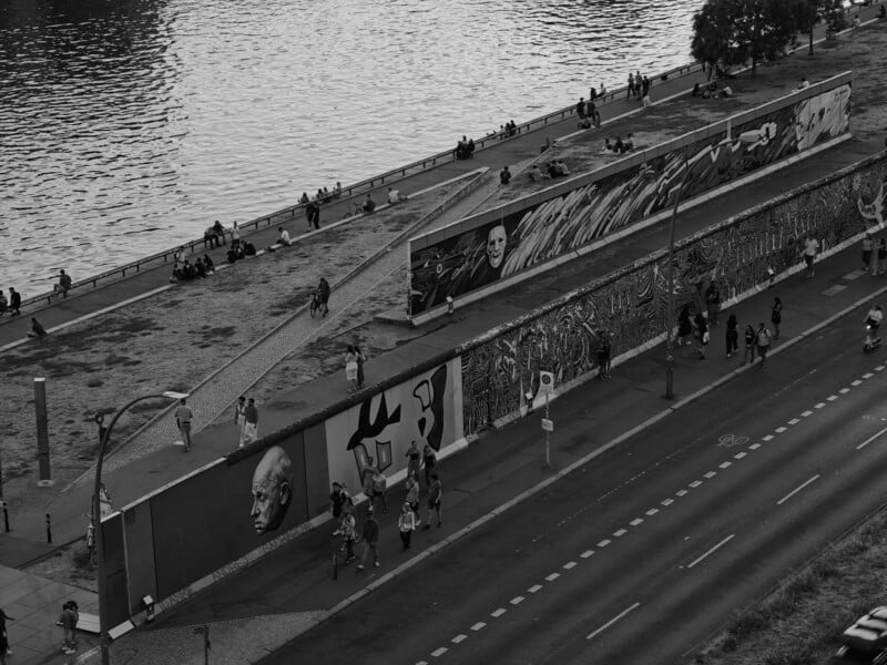 A black and white photo showing a riverside walkway with people walking and sitting. Large murals decorate the walls along the path, and a road with a bike lane runs nearby. Trees and the river add a natural backdrop.