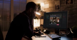 A person sits at a desk in a dimly lit room, looking at a computer monitor displaying a website with various image thumbnails. The room is softly illuminated by a desk lamp, creating a focused atmosphere.