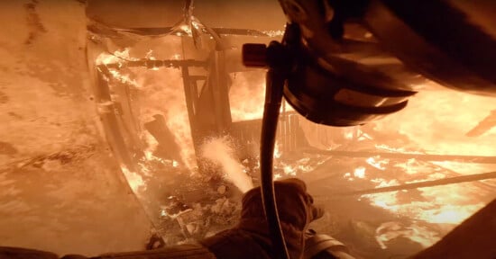 A firefighter battles intense flames inside a burning structure. The image shows the firefighter's perspective, with a gloved hand directing a hose at the fire. The walls and debris around are engulfed in bright orange flames.