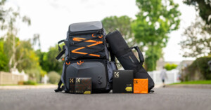 An assortment of outdoor gear is displayed on the road, including two black and orange backpacks, a black cylindrical bag, and three orange and black boxes with a logo and text. Background shows trees and houses, with a person walking in the distance.