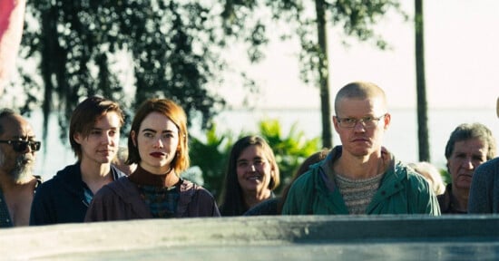 A group of people stand outdoors, facing the camera with neutral expressions. The setting is sunny and there are trees in the background. The group is diverse, including individuals with different hair colors and styles, with some wearing glasses.