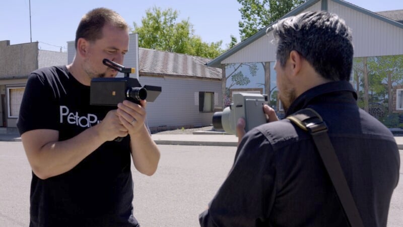 Two men facing each other outdoors, each holding a vintage camera up to their eyes. The man on the left is wearing a black T-shirt with white text, while the man on the right has a strap over his shoulder. They are near houses and trees with sunlight overhead.