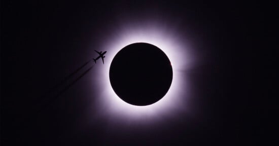 A plane is silhouetted against the dramatic backdrop of a solar eclipse, with the moon almost completely covering the sun, leaving only a bright halo of light. The sky around is dark, highlighting the striking contrast between the airplane and the eclipse.