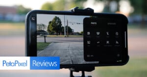 A smartphone mounted on a tripod, displaying a camera app interface with various settings visible. The phone screen shows a clear outdoor scene of a sidewalk, street, and trees in the background. "PetaPixel Reviews" text is overlayed at the bottom of the image.