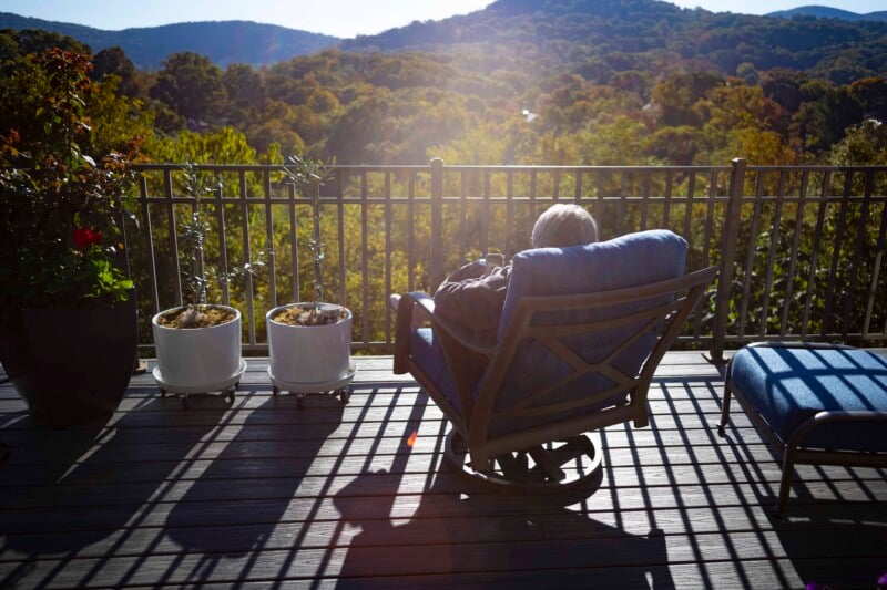 A person sitting in a cushioned chair on a wooden deck, facing away, with a scenic view of sunlit mountains and a forest. Two small potted plants and a blue cushioned bench are also on the deck.