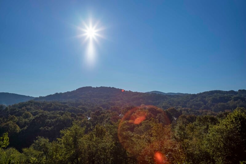 A bright sun shines over a lush green forest with hills in the background under a clear blue sky. Light lens flare is visible near the sun.