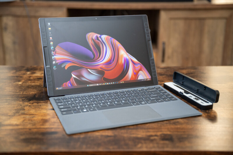 A laptop with a detachable keyboard is displayed on a wooden table. It has a colorful, abstract pattern on the screen. A pen is placed in a holder to the right of the laptop.