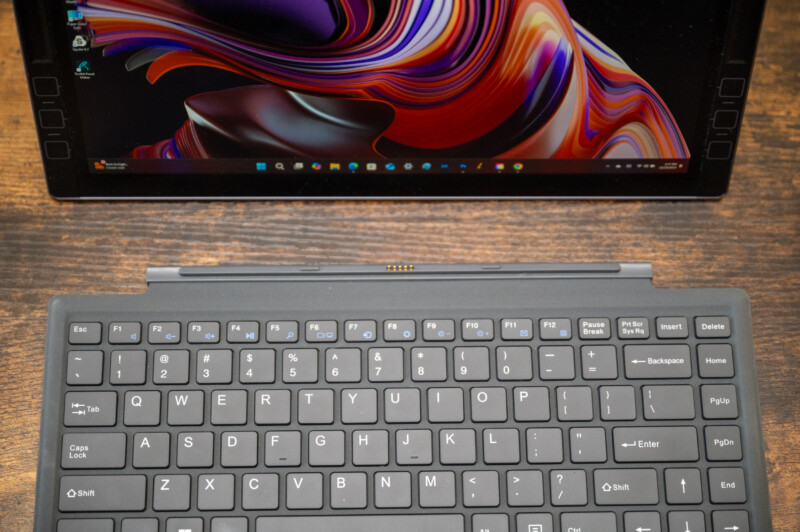 A laptop keyboard is attached to a tablet displaying a colorful, abstract wallpaper. The device is on a wooden surface, and the keyboard has various keys, including function keys, escape, and arrow keys. The screen shows part of the Windows taskbar.