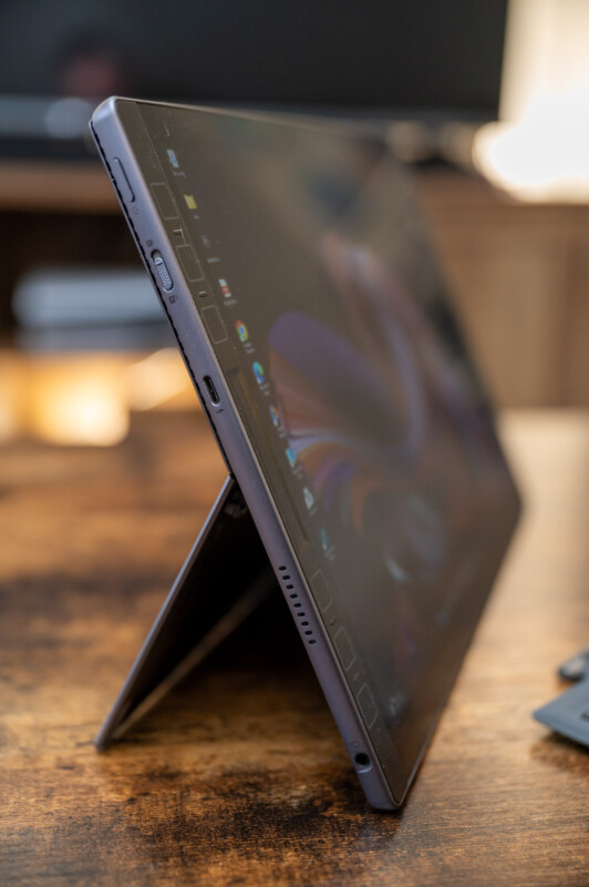 A side view of a laptop in a standing position, with its screen partially visible and displaying an abstract design. The device is placed on a wooden surface, with a blurred background highlighting its thin profile.