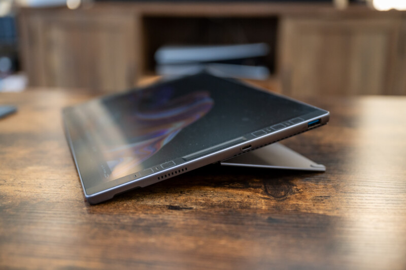 A thin, sleek tablet rests on a wooden surface, partially tilted upright with its stand extended. The screen is off, reflecting ambient light. Surrounding the tablet are unfocused wooden furniture elements in the background.