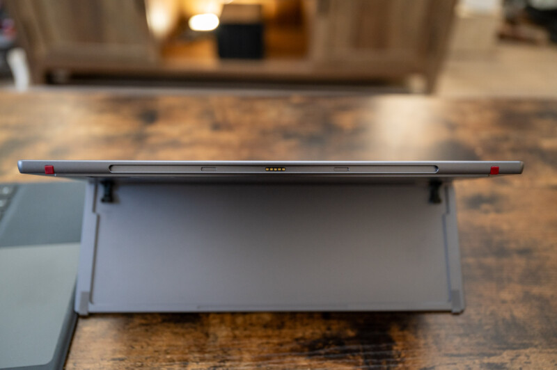 A close-up view of a tablet propped up on a stand, resting on a wooden table. The image shows the side profile of the tablet, highlighting its slim design and dark color, with a room softly lit in the background.