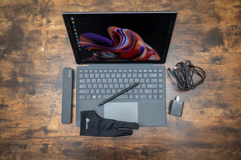 A digital drawing tablet with a vibrant screen is on a wooden table. Surrounding it are a stylus, a drawing glove, a pen case, a power adapter, and a USB cable. The screen displays colorful abstract art.