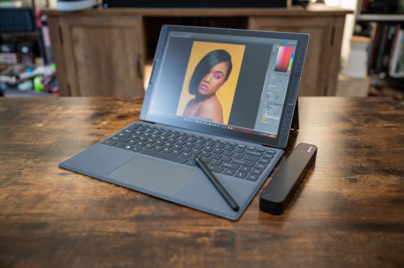 A tablet with a keyboard and stylus sits on a wooden table. An image editing software with a portrait is displayed on the screen. A wireless speaker is next to the tablet, and a wooden cabinet is in the background.